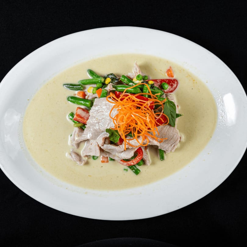 Thai green curry with sliced pork, green beans, bell peppers, and shredded carrots in a white bowl.