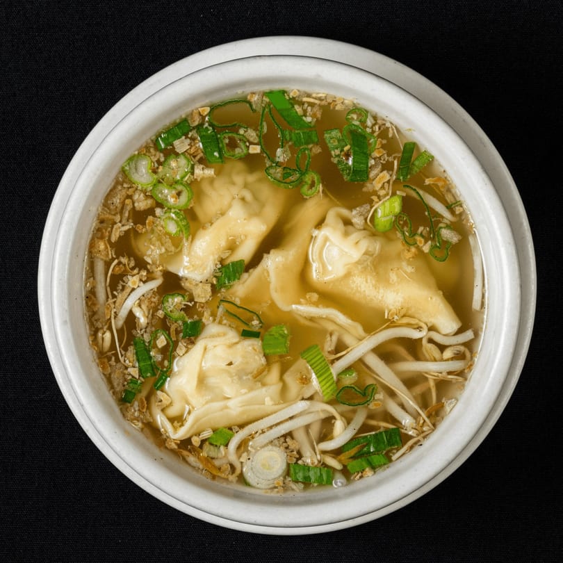 Overhead view of Thai wonton soup with bean sprouts, green onions, and fried garlic in a white bowl.