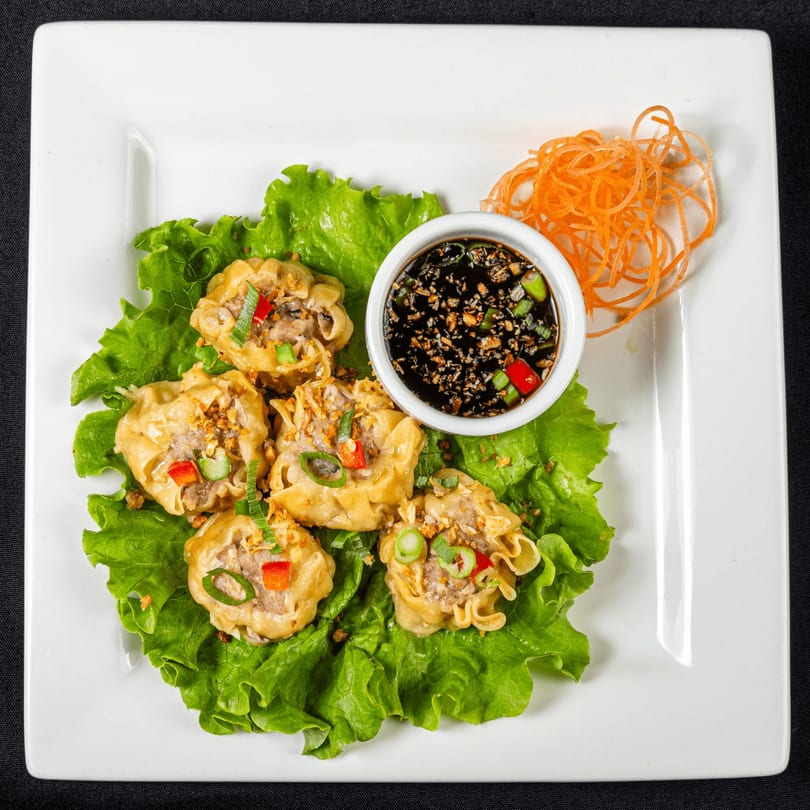 Thai Chilli Pepper's steamed dumplings with sauce and shredded carrots on a white plate.