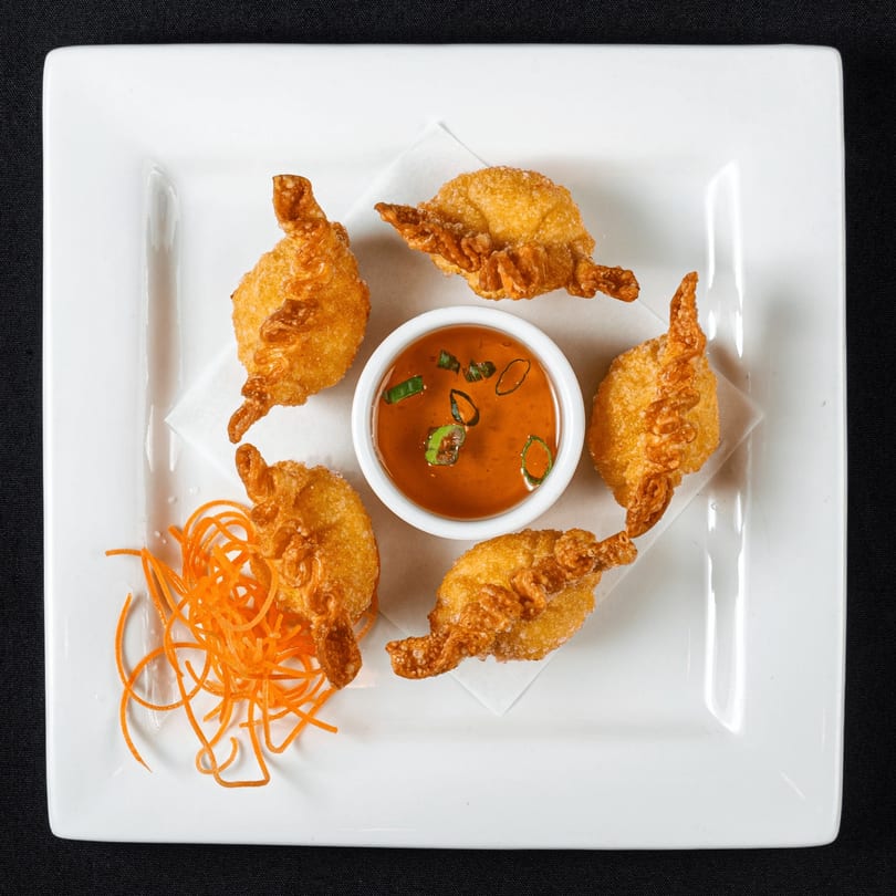 Five golden-fried Thai wontons arranged around a small bowl of chili dipping sauce and shredded carrots.